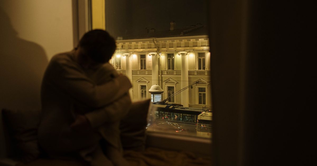 A pensive person sitting by the window at night overlooking a street with historical buildings.