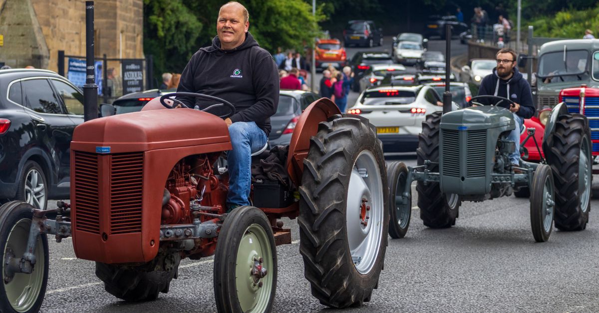 Two vintage tractors drive in a parade through Morpeth, England with spectators lining the street.